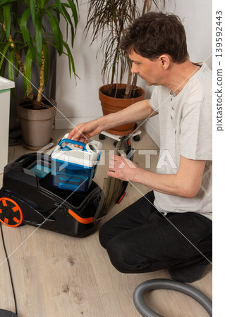 A 53-year-old man kneels on the floor in a room. He organizes attachments for a vacuum cleaner. Green plants and furniture are in the background as he gets ready to clean A 53-year-old man kneels on the floor in a room. He organizes attachments for a vacuum cleaner. Green plants and furniture are in the background as he gets ready to clean 139592443