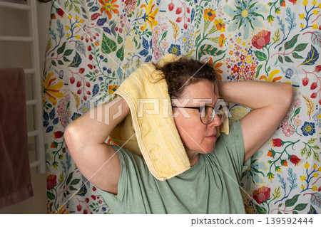 A 50-year-old woman starts her morning routine in the bathroom. She looks at herself in the mirror while holding a towel over her shoulder. The colorful background adds brightness 139592444