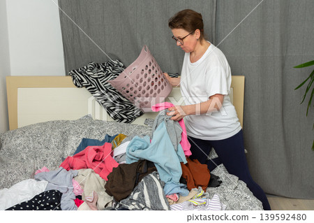 A 52-year-old woman pours clean laundry onto her bed. She is sorting clothes into different piles to organize them. This is happening inside her home in a relaxed setting A 52-year-old woman pours clean laundry onto her bed. She is sorting clothes into different piles to organize them. This is happening inside her home in a relaxed setting 139592480