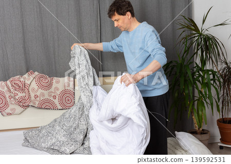 A 53-year-old man is in his bedroom changing bed linen. He holds a patterned bed cover and a white duvet as he prepares to make the bed clean and tidy 139592531