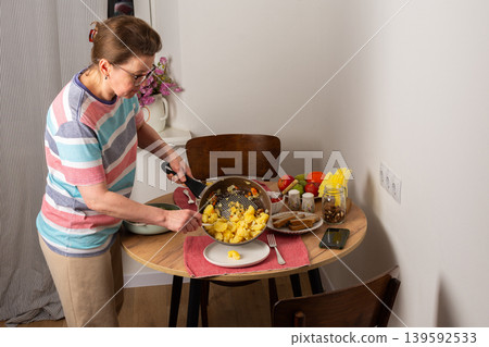 A 53-year-old woman prepares dinner at a round wooden table in her kitchen. She pours food from a colander onto a plate while arranging utensils and other items A 53-year-old woman prepares dinner at a round wooden table in her kitchen. She pours food from a colander onto a plate while arranging utensils and other items 139592533