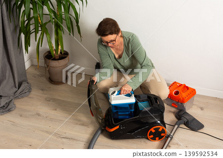Person getting ready to use a vacuum cleaner in a living room. The person adjusts the vacuum and prepares to clean the floor near a potted plant and cleaning tools Person getting ready to use a vacuum cleaner in a living room. The person adjusts the vacuum and prepares to clean the floor near a potted plant and cleaning tools 139592534