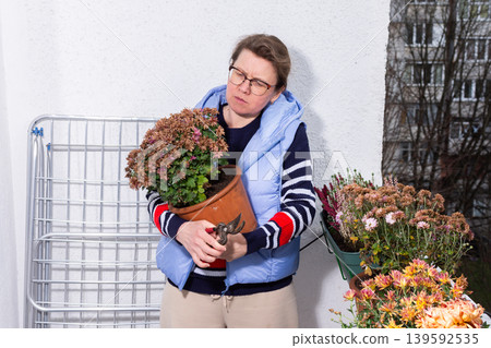 A 50-year-old woman works with autumn flowers on her balcony. She holds a flower pot, looking closely at the plants while surrounded by various blooms in pots A 50-year-old woman works with autumn flowers on her balcony. She holds a flower pot, looking closely at the plants while surrounded by various blooms in pots 139592535