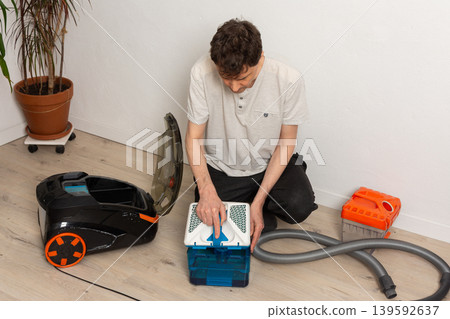 A 53-year-old man kneels on the floor as he prepares his vacuum cleaner for cleaning. He checks parts and organizes equipment in a well-lit room with plants 139592637