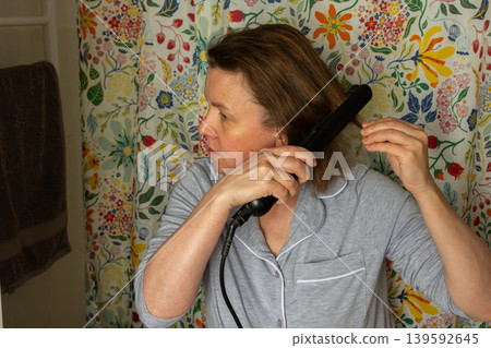 A 50-year-old woman styles her hair with a straightener in a bathroom. Floral curtains add color to the space. She is focused on her hair, preparing for the day ahead A 50-year-old woman styles her hair with a straightener in a bathroom. Floral curtains add color to the space. She is focused on her hair, preparing for the day ahead 139592645
