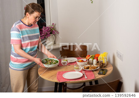 A 53-year-old woman sets the table for dinner. She carries a stack of plates and a glass of water. Fresh fruits and vegetables are arranged on the table, ready for the meal A 53-year-old woman sets the table for dinner. She carries a stack of plates and a glass of water. Fresh fruits and vegetables are arranged on the table, ready for the meal 139592646