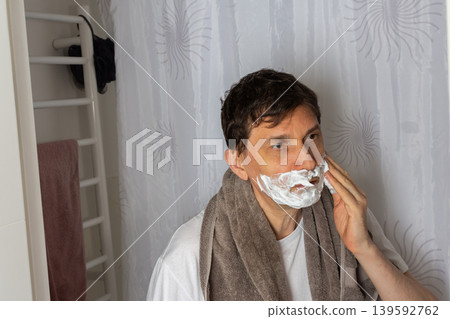 A 53-year-old man is in the bathroom applying shaving cream to his face while preparing for the day. He wears a towel around his neck and focuses on his grooming routine A 53-year-old man is in the bathroom applying shaving cream to his face while preparing for the day. He wears a towel around his neck and focuses on his grooming routine 139592762