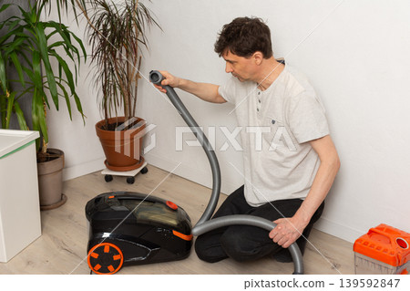 A man sits on the floor and connects the hose to a vacuum cleaner. He is focused on preparing the vacuum for cleaning. Indoor plants are nearby in the room 139592847