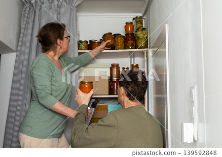 A married couple is busy placing canned goods on shelves in their pantry. They are in their city home, preparing for autumn. Boxes are organized, and the couple works together A married couple is busy placing canned goods on shelves in their pantry. They are in their city home, preparing for autumn. Boxes are organized, and the couple works together 139592848