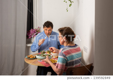 A married couple is having dinner together at home. They sit at a small table near a window with a bowl of salad and other food. The light is warm and cozy in the evening A married couple is having dinner together at home. They sit at a small table near a window with a bowl of salad and other food. The light is warm and cozy in the evening 139592849