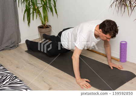 A man, 53 years old, lies on a yoga mat at home, performing a stretching exercise. He focuses on his fitness routine in a simple indoor space with plants around A man, 53 years old, lies on a yoga mat at home, performing a stretching exercise. He focuses on his fitness routine in a simple indoor space with plants around 139592850