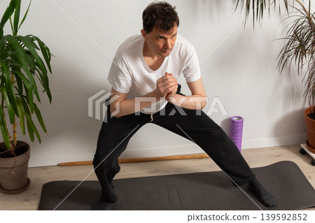 A 53-year-old man practices exercises at home on a yoga mat. He focuses on his strength and flexibility while avoiding distractions from the surroundings. Plants stand around him A 53-year-old man practices exercises at home on a yoga mat. He focuses on his strength and flexibility while avoiding distractions from the surroundings. Plants stand around him 139592852