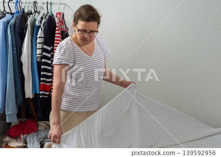 A 53-year-old woman is working in her bedroom. She is changing the bed linen and placing new sheets on the bed. Clothes are seen nearby in the room A 53-year-old woman is working in her bedroom. She is changing the bed linen and placing new sheets on the bed. Clothes are seen nearby in the room 139592956