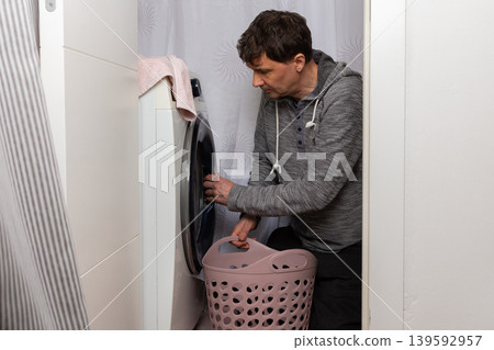 A man is taking clothes out of a washing machine. He is putting them into a basket. This is happening in a laundry room at home. The lighting shows it is daytime A man is taking clothes out of a washing machine. He is putting them into a basket. This is happening in a laundry room at home. The lighting shows it is daytime 139592957