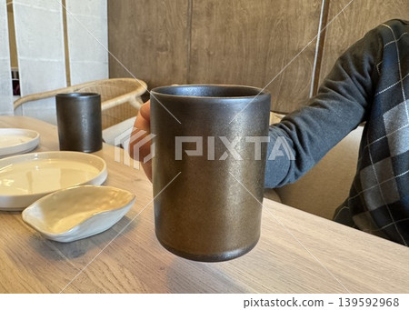 Hand holding dark ceramic coffee mug at wooden table, cozy cafe interior with modern tableware close-up Hand holding dark ceramic coffee mug at wooden table, cozy cafe interior with modern tableware close-up 139592968