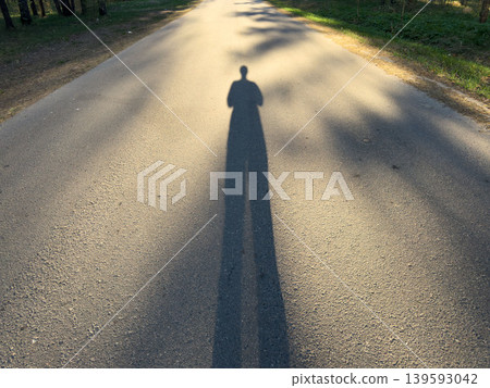 A long human shadow on a deserted asphalt road at sunset, a silhouette reflection in a forest park in the golden evening light A long human shadow on a deserted asphalt road at sunset, a silhouette reflection in a forest park in the golden evening light 139593042