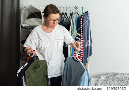 Woman sorts and hangs washed clothes on a hanger. She holds a blue sweatshirt and several other items in her hands. This scene takes place in a home environment Woman sorts and hangs washed clothes on a hanger. She holds a blue sweatshirt and several other items in her hands. This scene takes place in a home environment 139593085