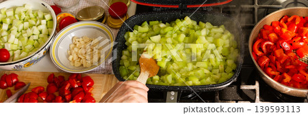 A woman stirs green squash in a skillet while preparing garlic and chopped vegetables on a nearby table. She is getting ready to make squash caviar for winter storage, banner A woman stirs green squash in a skillet while preparing garlic and chopped vegetables on a nearby table. She is getting ready to make squash caviar for winter storage, banner 139593113