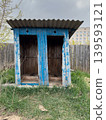 An abandoned blue wooden outbuilding with a corrugated roof, an old outdoor toilet in an overgrown yard in a rural area 139593121