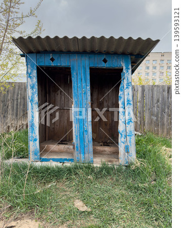 An abandoned blue wooden outbuilding with a corrugated roof, an old outdoor toilet in an overgrown yard in a rural area An abandoned blue wooden outbuilding with a corrugated roof, an old outdoor toilet in an overgrown yard in a rural area 139593121