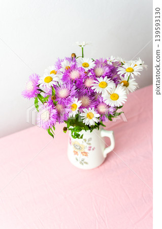 A colorful bouquet of daisies and purple flowers arranged in a charming pitcher rests on a pink tablecloth, bringing joy to the soft, simple setting A colorful bouquet of daisies and purple flowers arranged in a charming pitcher rests on a pink tablecloth, bringing joy to the soft, simple setting 139593130