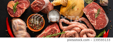 Different types of meat are arranged on a dark surface. The setup includes beef steaks, chicken legs, quail, pork sausages, and seasonings like salt and herbs, banner Different types of meat are arranged on a dark surface. The setup includes beef steaks, chicken legs, quail, pork sausages, and seasonings like salt and herbs, banner 139593133