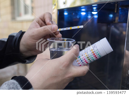 Man checking water quality at a public vending machine using a test strip. Analysis of purified drinking water in a plastic cup. Safety, pH, and hardness control of urban water supply. 139593477