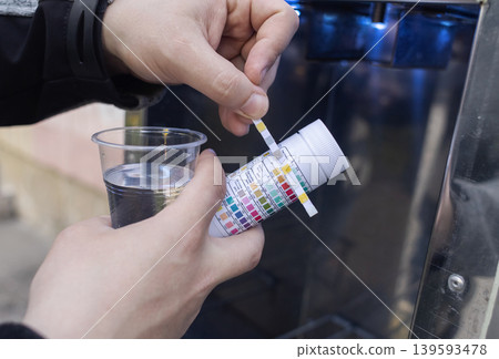 Man checking water quality at a public vending machine using a test strip. Analysis of purified drinking water in a plastic cup. Safety, pH, and hardness control of urban water supply. 139593478