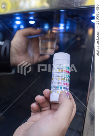 Man checking water quality at a public vending machine using a test strip. Analysis of purified drinking water in a plastic cup. Safety, pH, and hardness control of urban water supply. 139593480