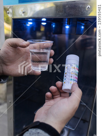 Man checking water quality at a public vending machine using a test strip. Analysis of purified drinking water in a plastic cup. Safety, pH, and hardness control of urban water supply. 139593481