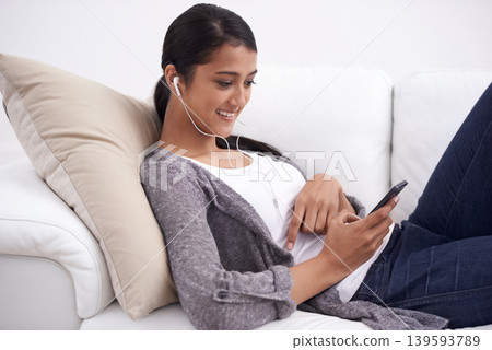 Young woman, smartphone and earphones, relax on couch with social media, communication and music. Listening to radio, podcast or watch video online, website or blog with mobile app and tech at home Young woman, smartphone and earphones, relax on couch with social media, communication and music. Listening to radio, podcast or watch video online, website or blog with mobile app and tech at home 139593789
