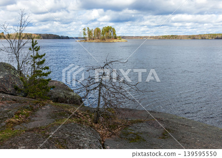 A quiet rocky shoreline looks out across a calm lake to a small tree-covered island A quiet rocky shoreline looks out across a calm lake to a small tree-covered island 139595550