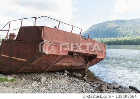 A rusty abandoned boat sits on a rocky lake shore beneath forested mountains A rusty abandoned boat sits on a rocky lake shore beneath forested mountains 139595564