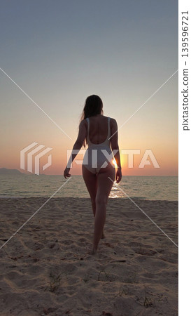 Woman in white swimsuit walking on beach to sunset over sea Woman in white swimsuit walking on beach to sunset over sea 139596721