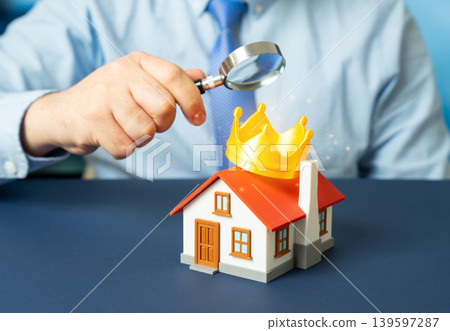 A man examines a house with a golden crown under a magnifying glass. Evaluating premium housing. Desirable property qualities and real estate ranking. A man examines a house with a golden crown under a magnifying glass. Evaluating premium housing. Desirable property qualities and real estate ranking. 139597287