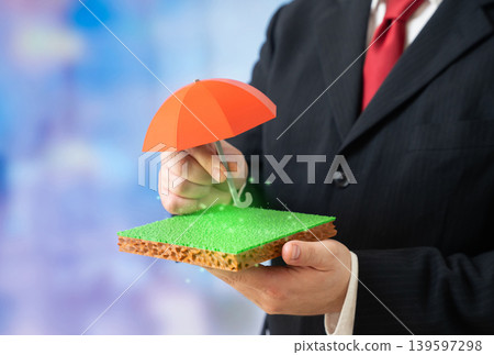 A man holds an umbrella over a plot of land. Insurance and protection of land rights. Legal guarantees for investors. Property risk management. A man holds an umbrella over a plot of land. Insurance and protection of land rights. Legal guarantees for investors. Property risk management. 139597298