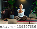 Middle aged woman practicing online yoga workout using laptop in modern Scandi dark interior home. Healthy Lifestyle. Sport activity for mature wellbeing. Fitness online training. Mindful movement 139597636