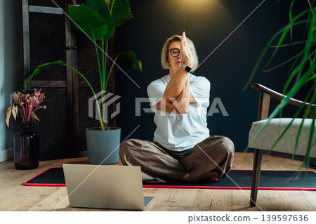 Middle aged woman practicing online yoga workout using laptop in modern Scandi dark interior home. Healthy Lifestyle. Sport activity for mature wellbeing. Fitness online training. Mindful movement 139597636