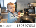 Baby boy standing on learning tower near induction stove with frying pan and holding a spatula in hand. Toddler repeating actions after parents, learning to cook food. Everyday kid development routine 139597638