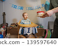 Baby boy sits in high chair looking surprised while father holds a birthday cake with candles. Colorful decorations fill the living room. Family gathers to celebrate first birthday occasion. 139597640