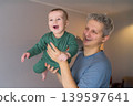 Father and toddler son having fun. Young father lifts his joyful baby in a cozy room. Baby laughing, showing delight. They enjoy their time together at home. Dad playing with baby, happy childhood. 139597641