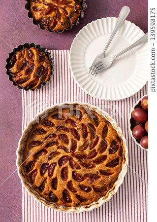 A plum pie is placed on a table with thinly sliced plums visible on top. Bowls of fresh plums are nearby with empty plates waiting for dessert A plum pie is placed on a table with thinly sliced plums visible on top. Bowls of fresh plums are nearby with empty plates waiting for dessert 139598175