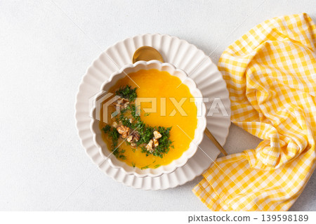 A bowl of pumpkin cream soup sits on a plate. The soup contains nuts and herbs. A yellow checkered napkin lies nearby. This meal can be enjoyed at breakfast or lunch 139598189
