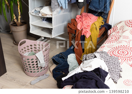 Clothes are scattered on the floor and inside a basket in a room. A chair holds additional clothes. The scene shows items ready for washing after daily use and activities Clothes are scattered on the floor and inside a basket in a room. A chair holds additional clothes. The scene shows items ready for washing after daily use and activities 139598210