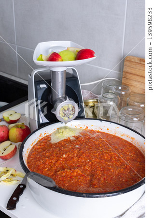 A person is using a grinder to prepare Adjika, a Georgian spicy seasoning, in a kitchen. The bowl contains the mixture while apples and jars are nearby on the counter A person is using a grinder to prepare Adjika, a Georgian spicy seasoning, in a kitchen. The bowl contains the mixture while apples and jars are nearby on the counter 139598233
