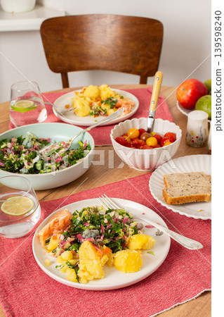 Boiled potatoes and salmon are served alongside a fresh salad made of greens, purple cauliflower, and pink radish. Whole grain bread and cherry tomatoes are also present on the table 139598240