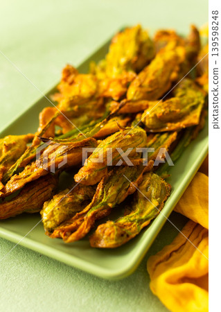 Delicious deep-fried zucchini flowers are neatly arranged on a green platter, showcasing their crispy texture. A bright yellow napkin adds warmth to the scene 139598248