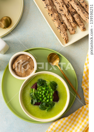 A creamy soup made from broccoli and green peas is served alongside liver pate and grissini sticks on a simple table setting. Fresh ingredients are arranged neatly for a healthy meal A creamy soup made from broccoli and green peas is served alongside liver pate and grissini sticks on a simple table setting. Fresh ingredients are arranged neatly for a healthy meal 139598251