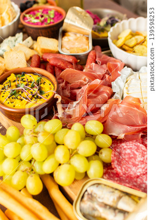 Gathered around a beautifully arranged charcuterie board filled with assorted cheeses, delicious sausages, creamy hummus, and fresh grapes. Perfect for a small gathering Gathered around a beautifully arranged charcuterie board filled with assorted cheeses, delicious sausages, creamy hummus, and fresh grapes. Perfect for a small gathering 139598253