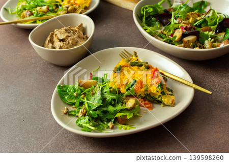 A plate features baked eggs with vegetables alongside a fresh arugula salad. Small bowls hold liver pate and baked pear pieces. This meal is set on a neutral table A plate features baked eggs with vegetables alongside a fresh arugula salad. Small bowls hold liver pate and baked pear pieces. This meal is set on a neutral table 139598260
